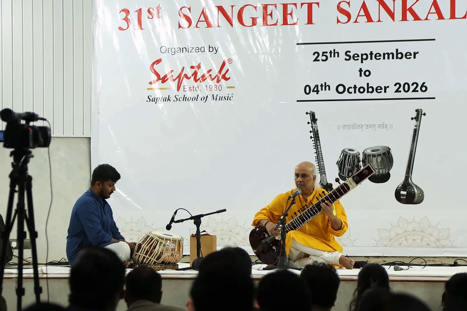 View from the back of the hall over the audience, with a video camera on a tripod and the sitar and tabla duo on stage beneath the Saptak banner