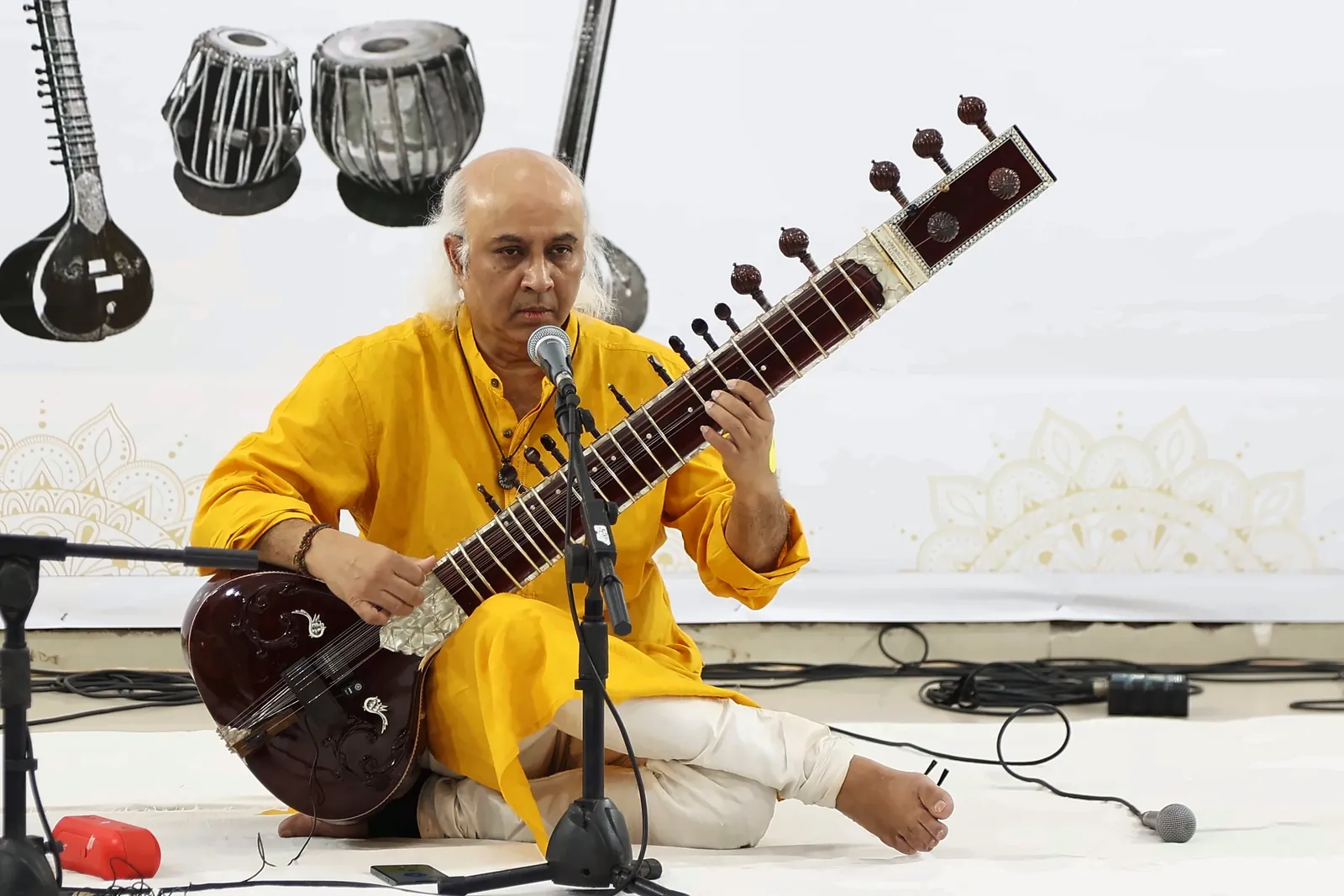 Close-up of Vatsal Dave playing sitar into a microphone, eyes lowered to the frets, with sitar and tabla artwork on the backdrop