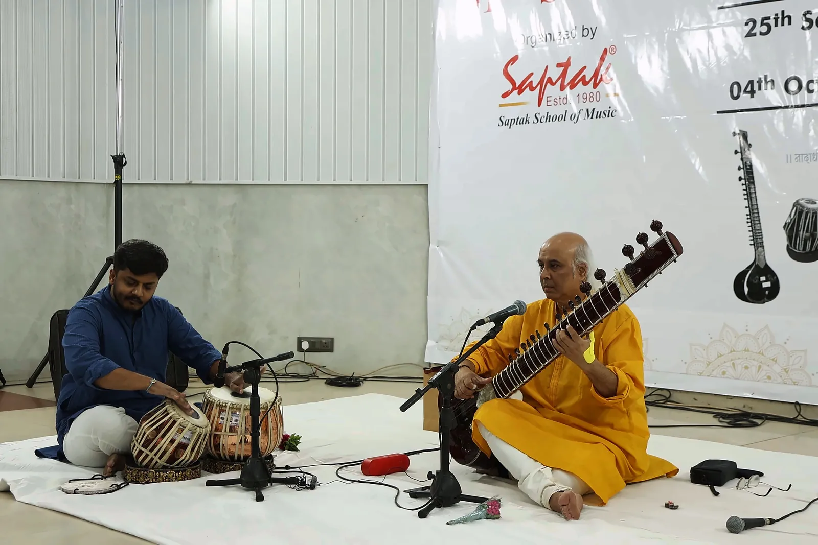 Side view of the stage with Riddhish Vora playing tabla on the left and Vatsal Dave playing sitar on the right, part of the Saptak banner visible